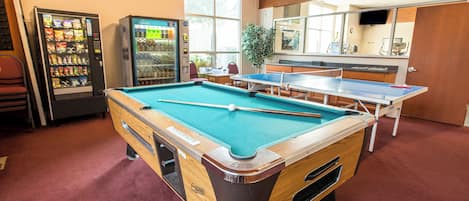A pool table with a blue felt surface, a ping pong table, and vending machines in a room with red carpet flooring.