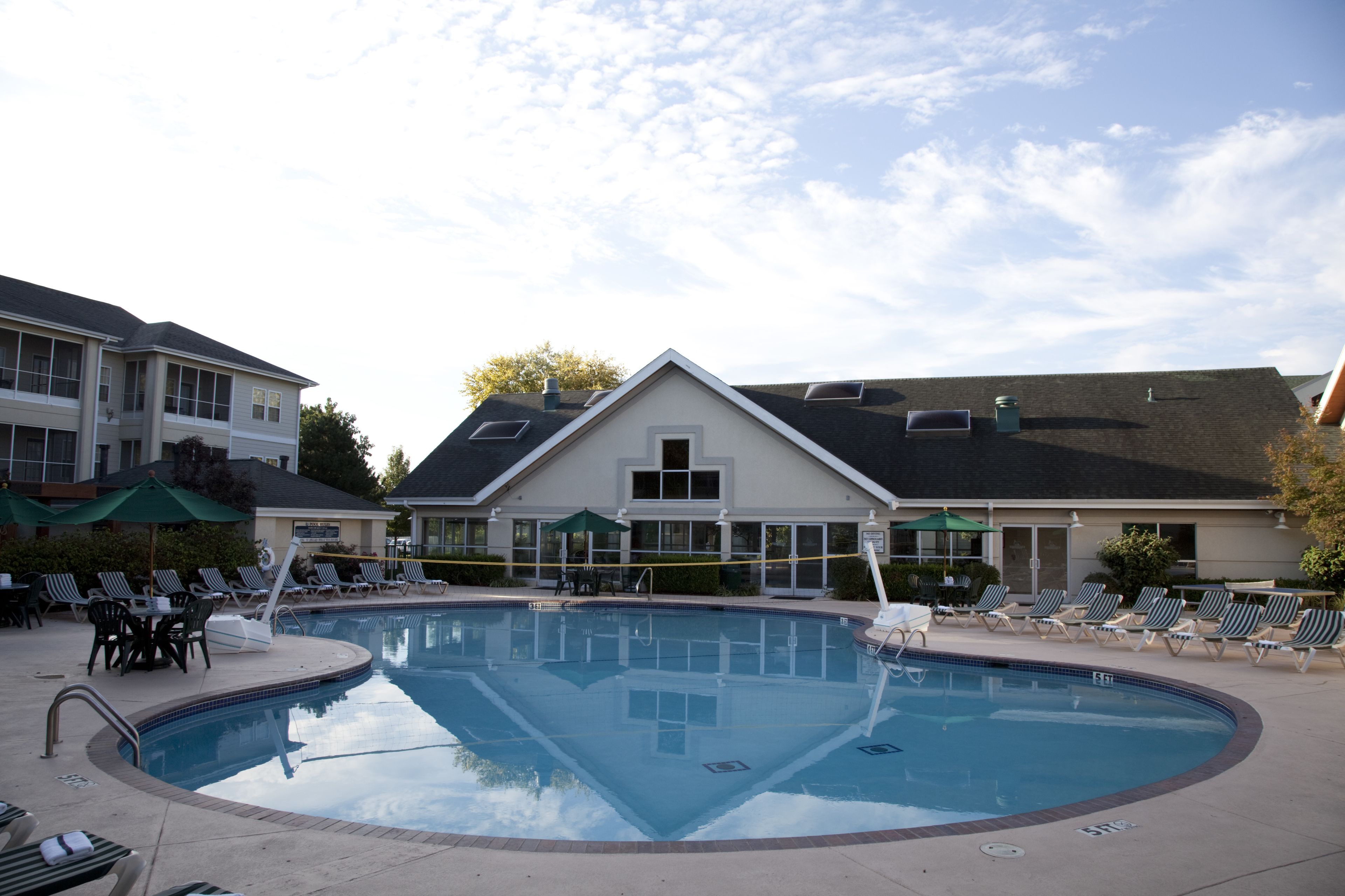 Ein Schwimmbecken mit Liegestühlen und Sonnenschirmen, umgeben von Gebäuden mit großen Fenstern und einer überdachten Terrasse.