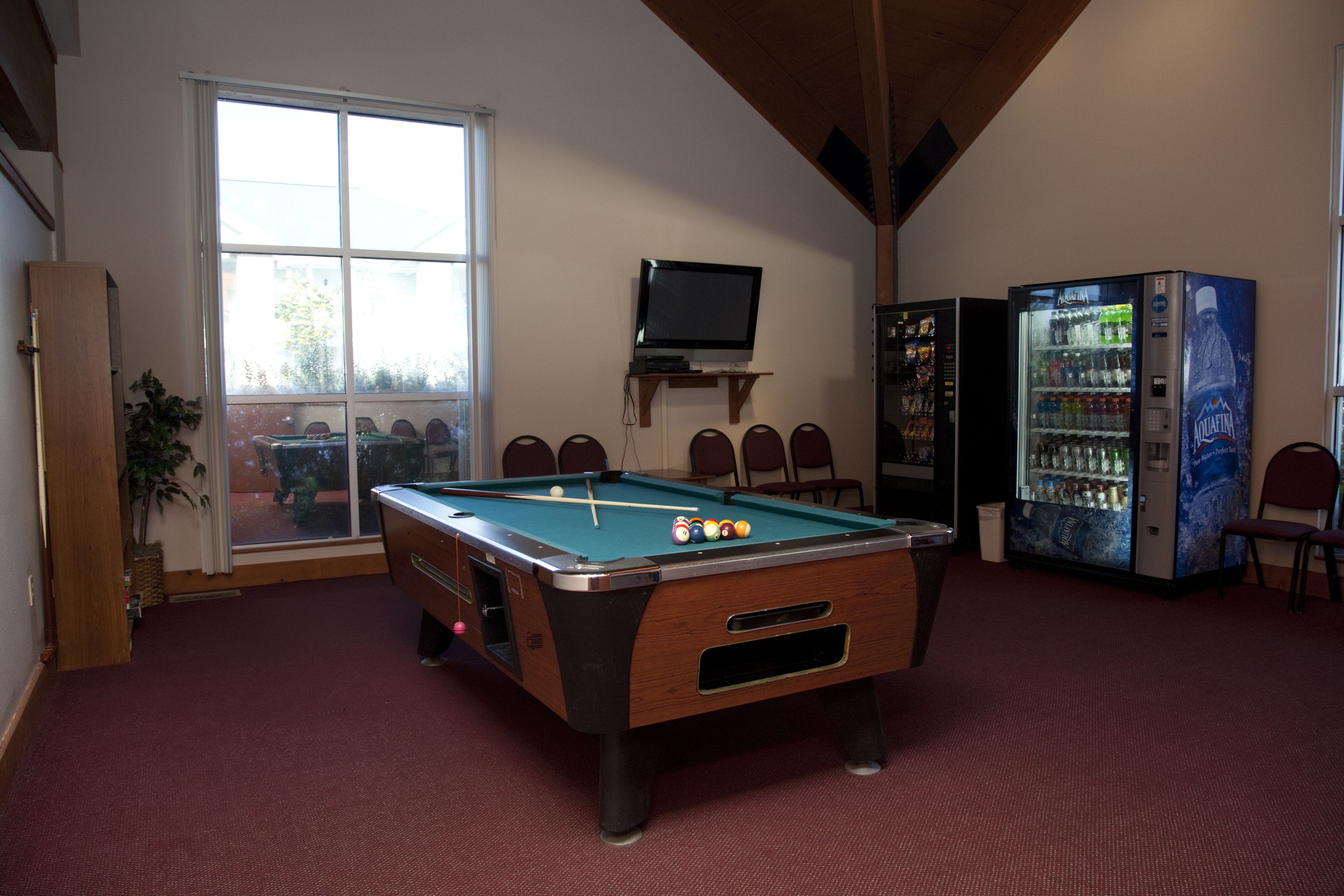 A pool table in a room with a large window, a television on a shelf, and a vending machine.