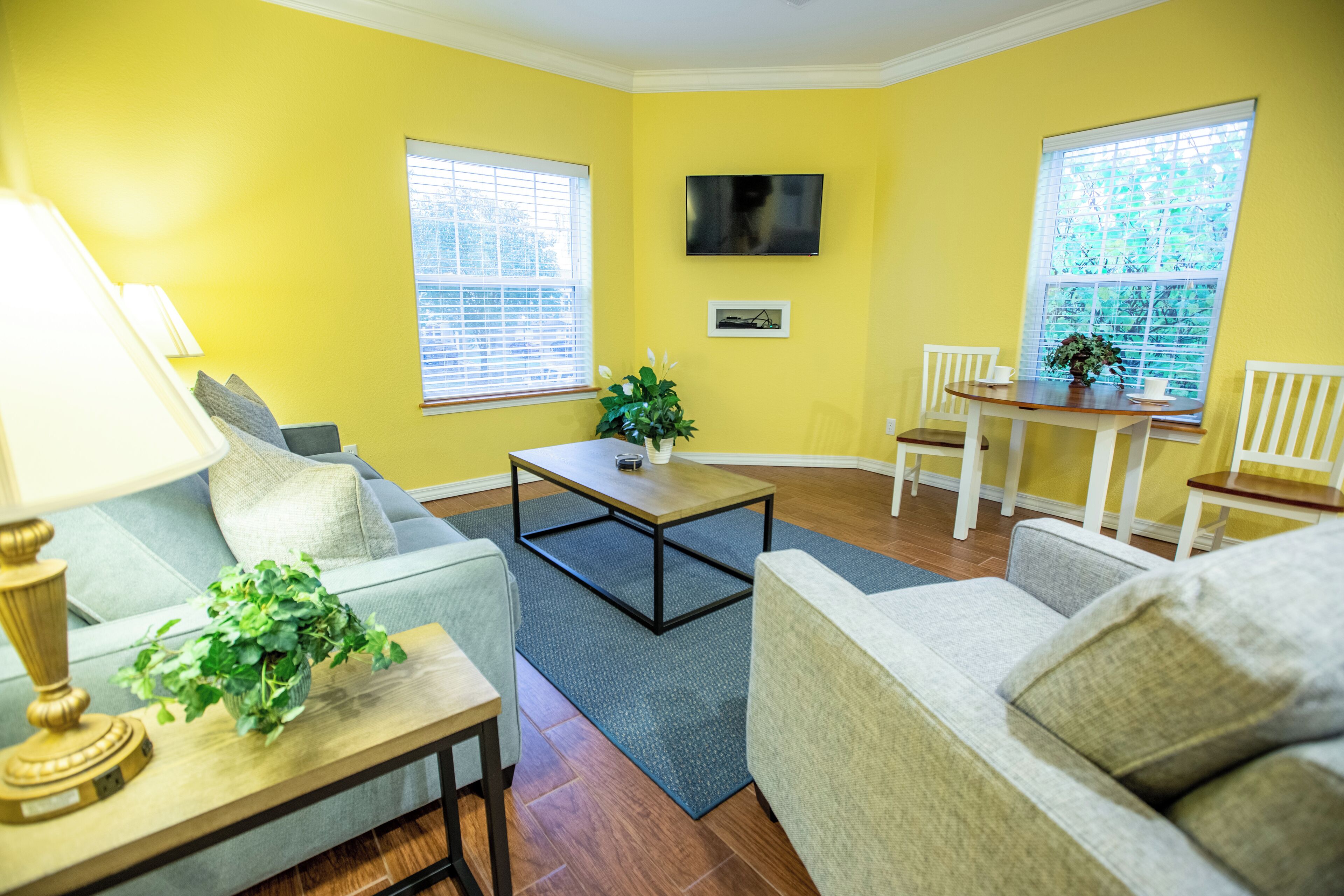 A living room with yellow walls, a TV mounted on the wall, a wooden coffee table, a sofa, a dining table with chairs, and a window with blinds.