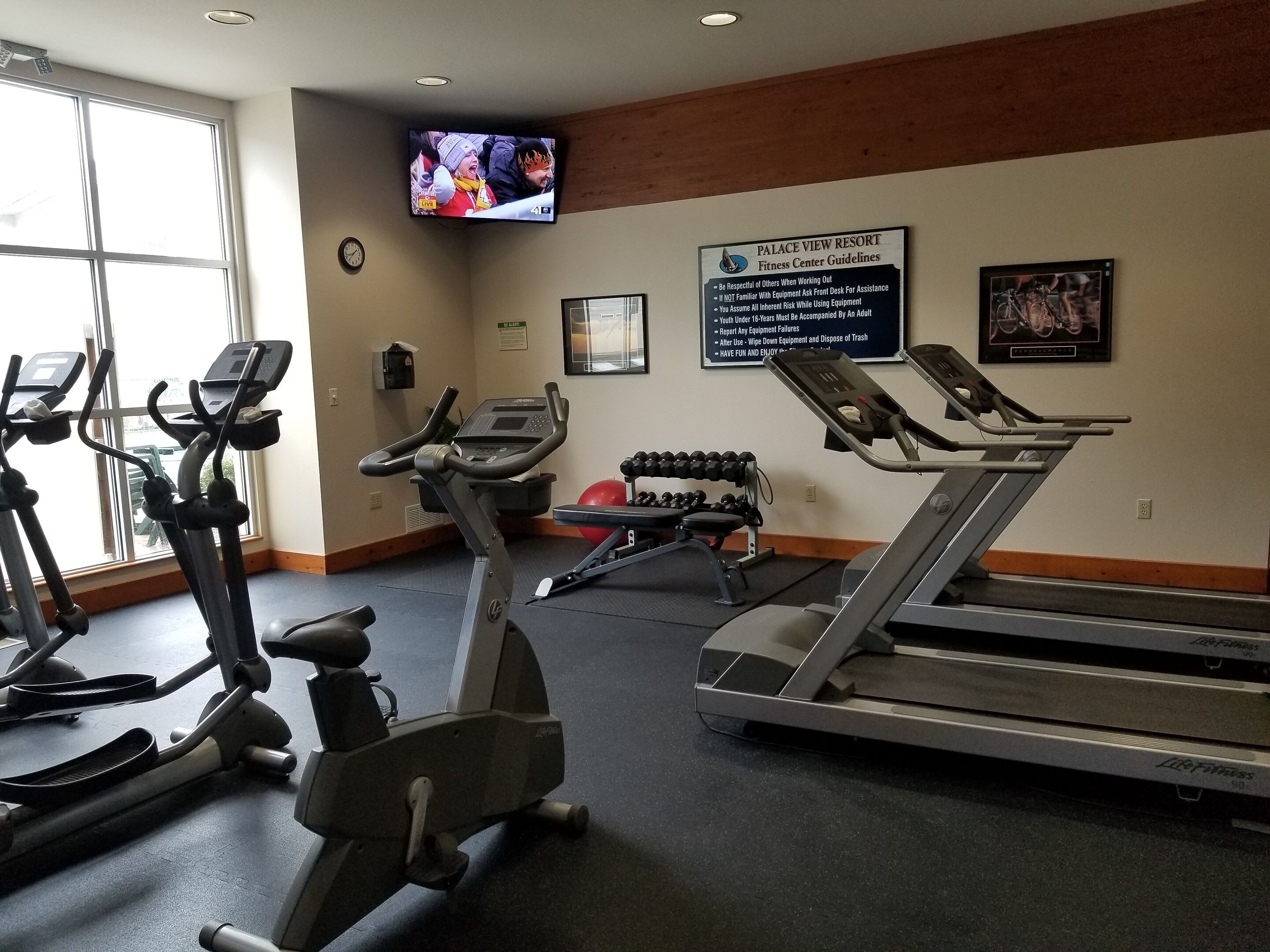 Un gimnasio con cintas de correr, elípticas y mancuernas. Un televisor en la pared muestra un evento deportivo.