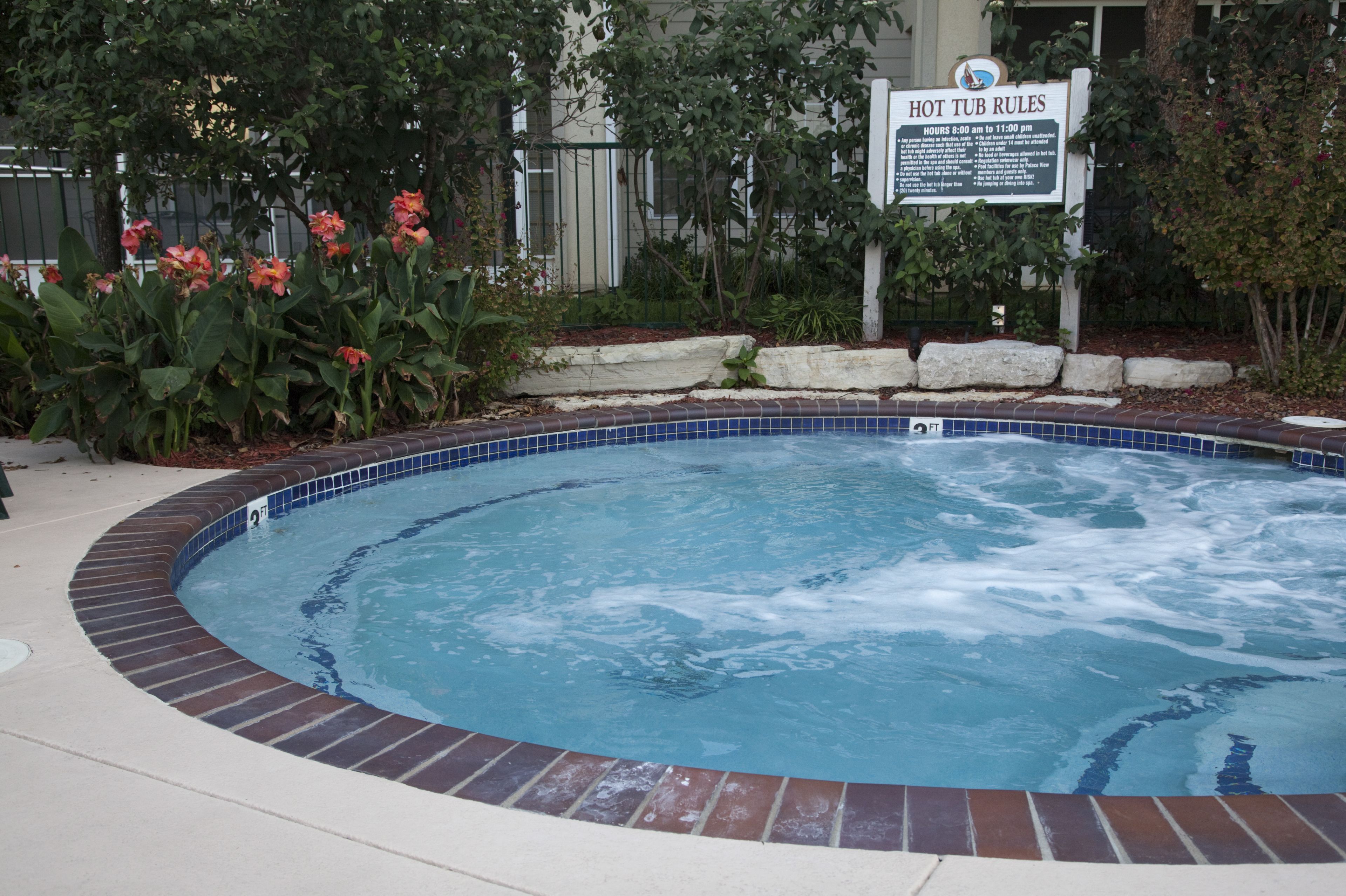 A hot tub with a sign detailing hot tub rules, surrounded by a brick pool edge and lush greenery.