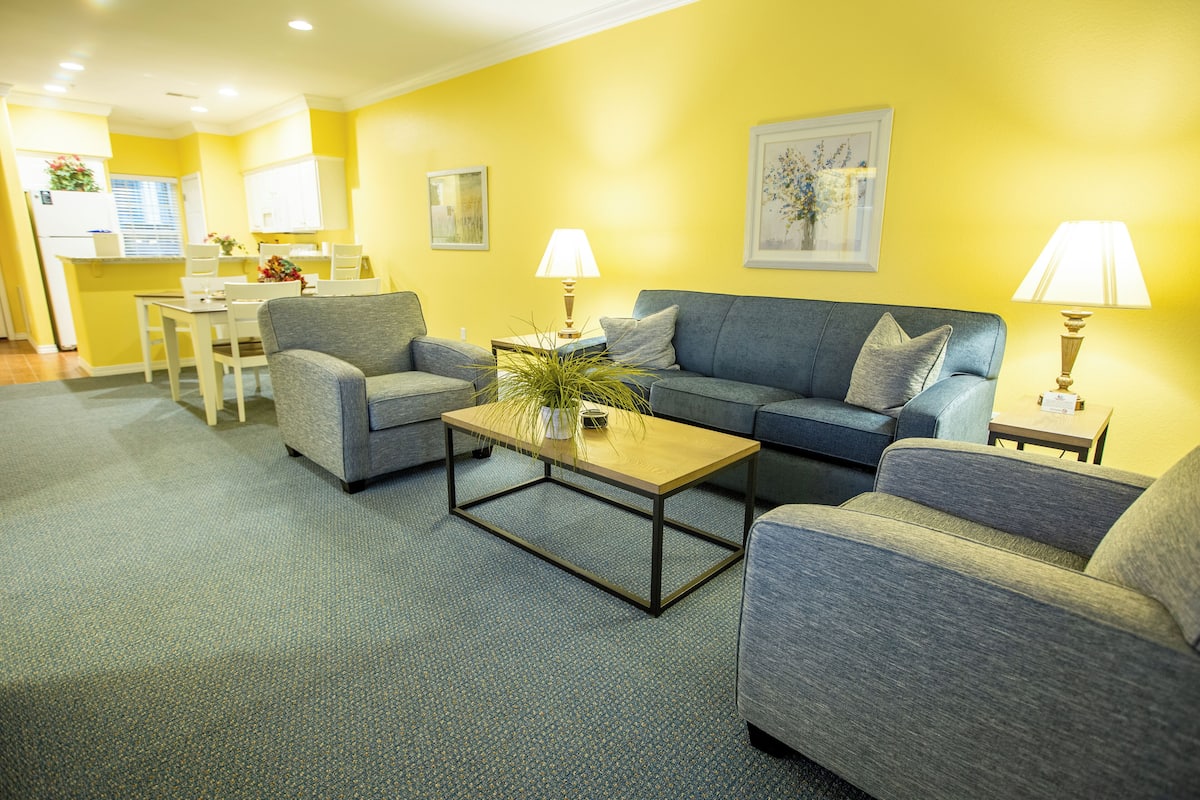 A living room with a sofa, two armchairs, a coffee table, and a lamp. There is a dining table with chairs and a kitchen area in the background.