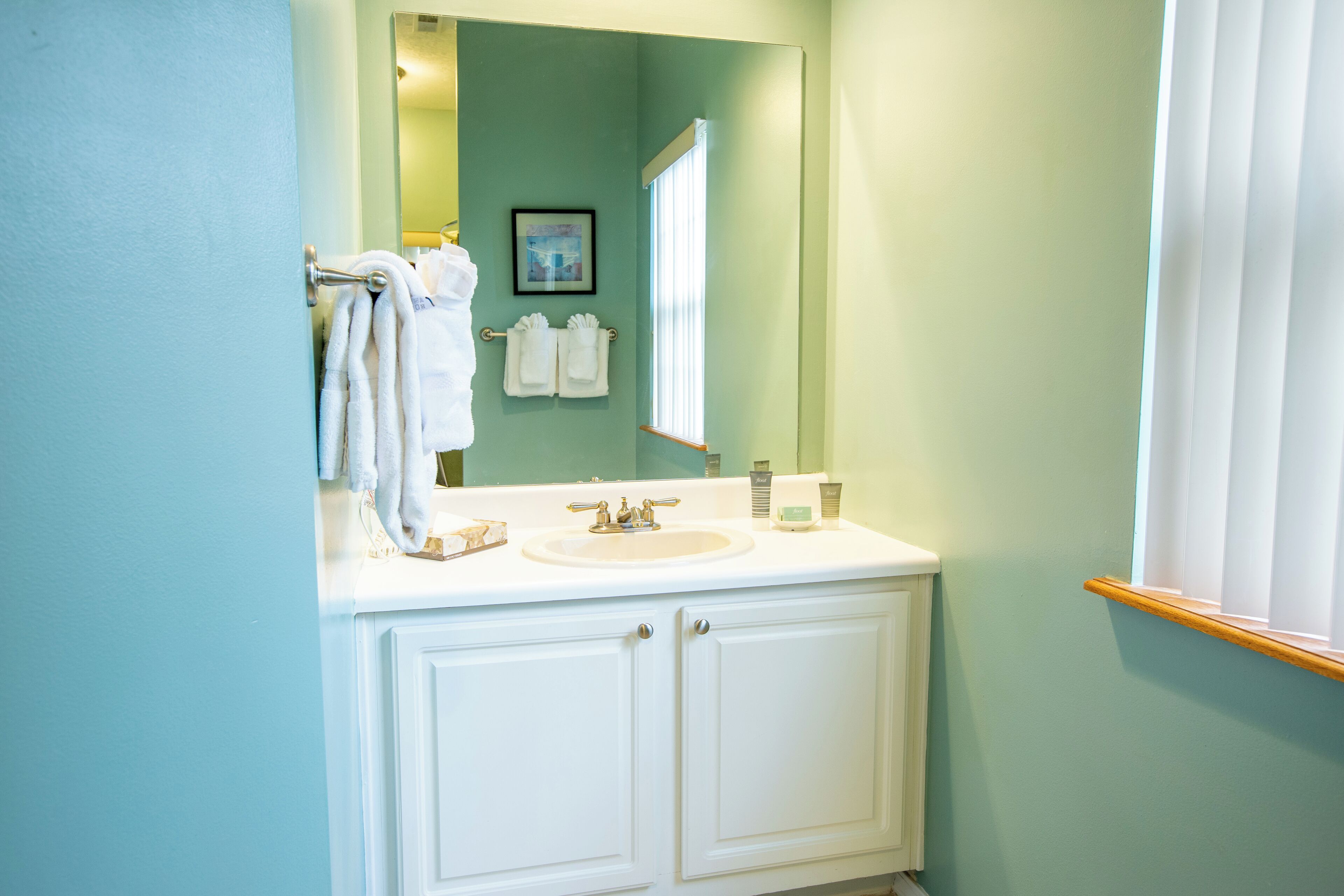 Une salle de bain avec un meuble-lavabo blanc, un évier et un miroir. Des serviettes sont suspendues sur un porte-serviettes et un tableau encadré est accroché au mur.