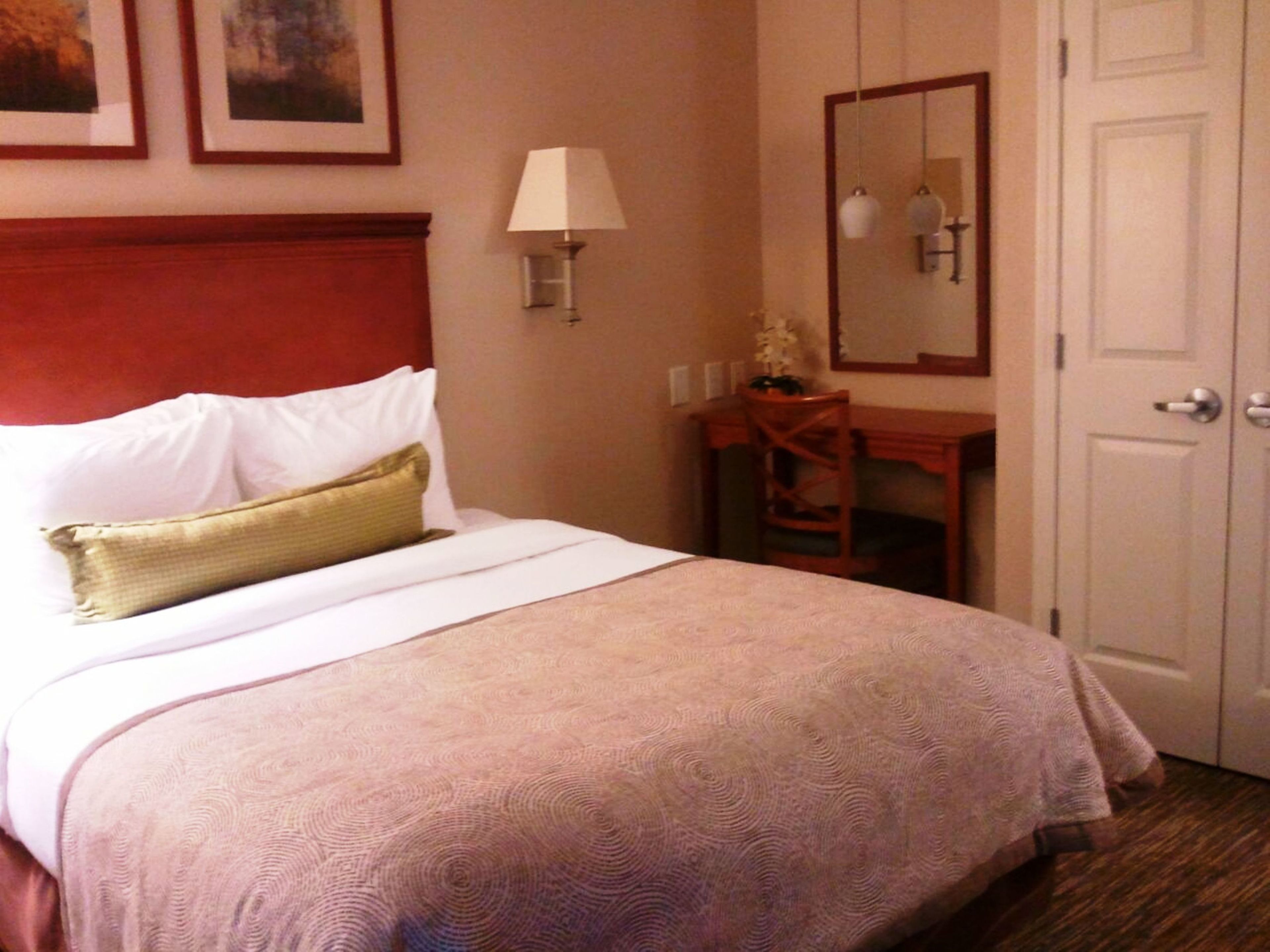 A hotel room with a bed, a desk, a chair, a lamp, and a door.