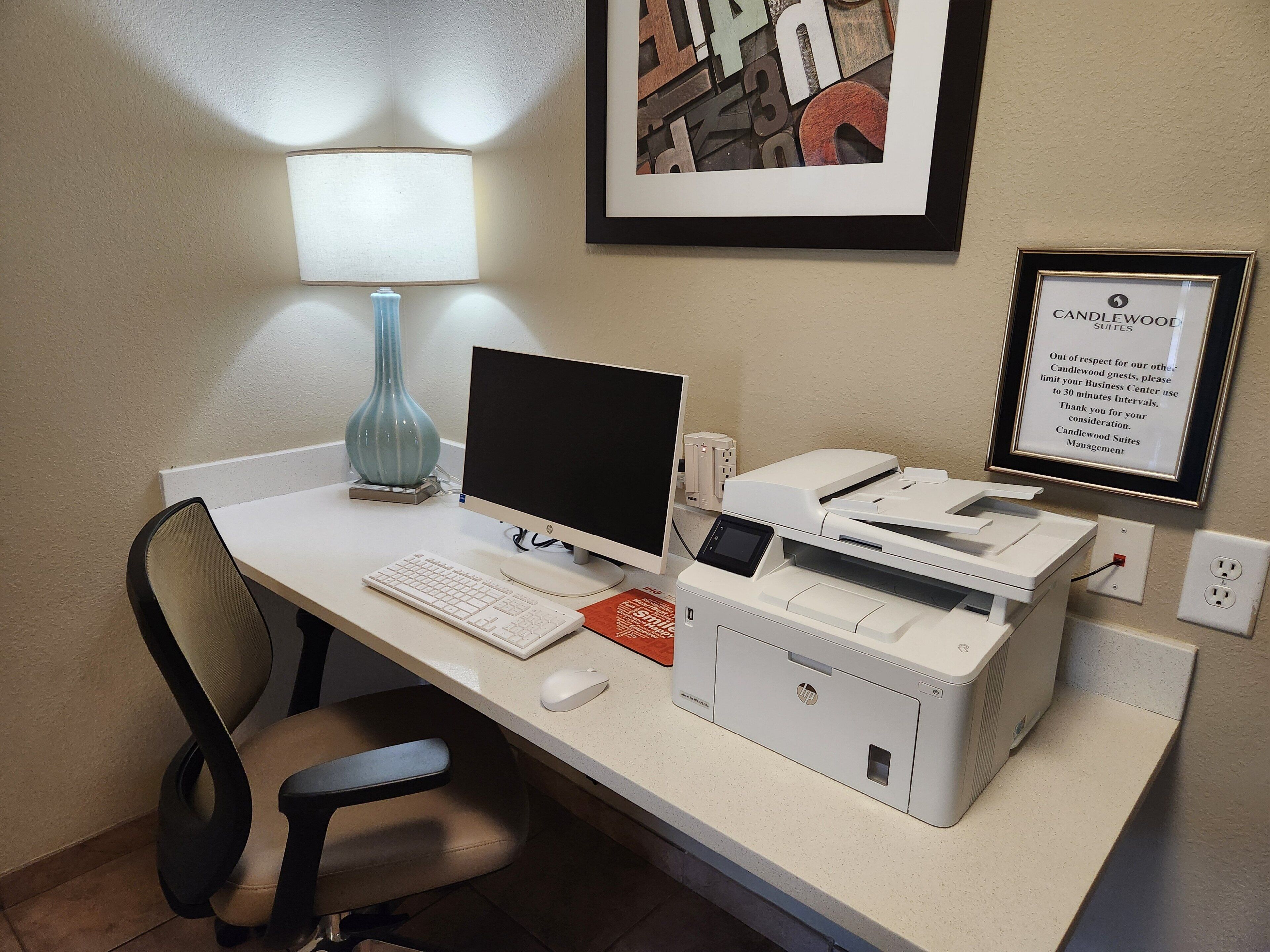 Een hotel werkplek met een bureau, computer, printer, stoel, lamp en een lijst in een lijstwerk.