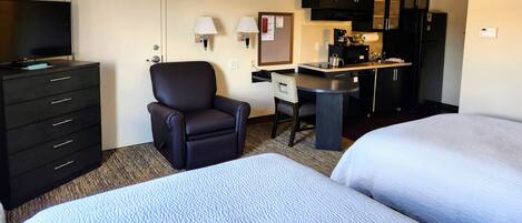 A hotel room with two beds, a desk with a chair, a television, a minibar, and a kitchenette.