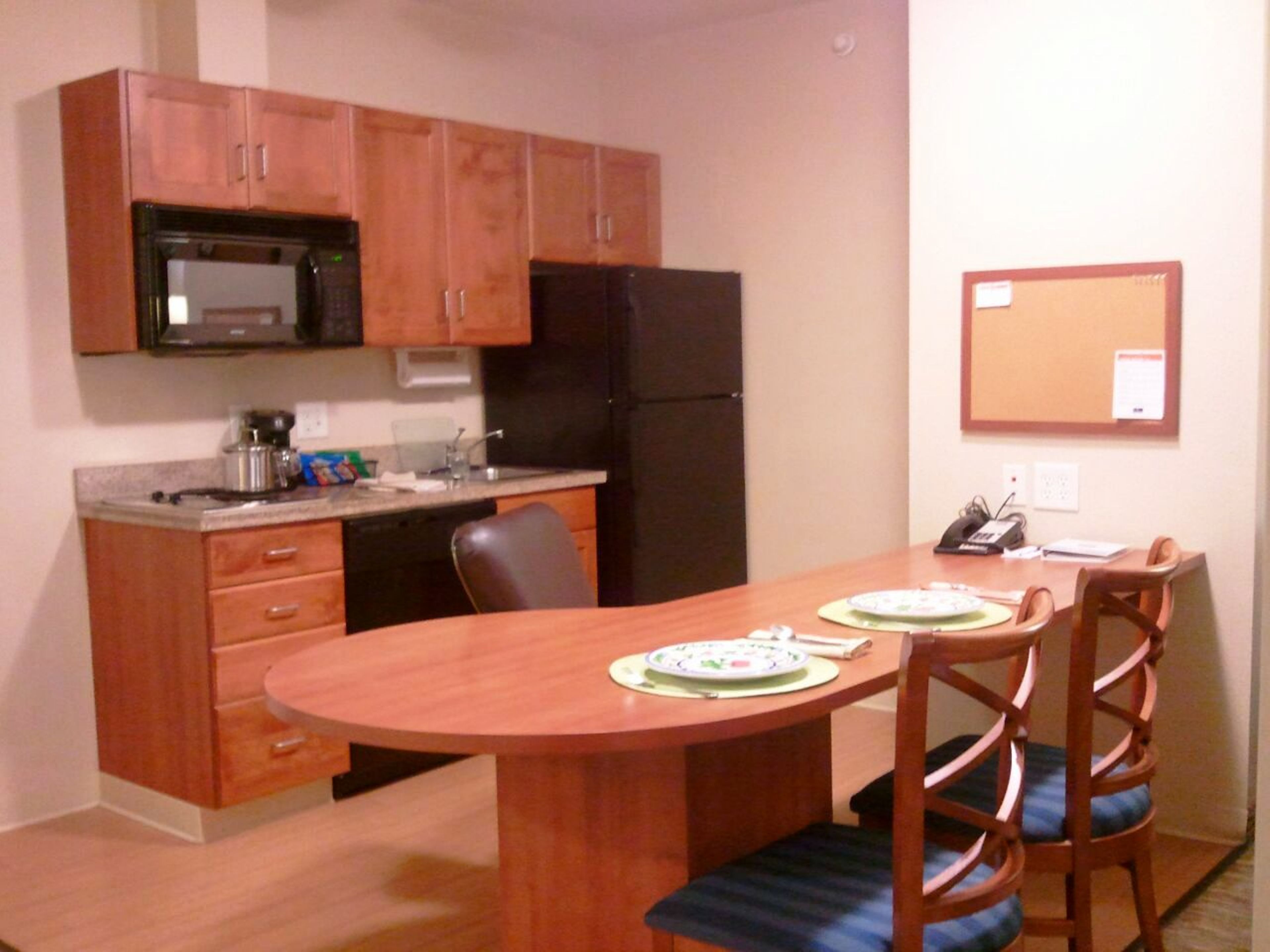 Una cocina compacta con armarios de madera, un refrigerador negro, un microondas y un lavamanos. Hay una mesa de comedor con sillas y un tablón de anuncios en la pared.