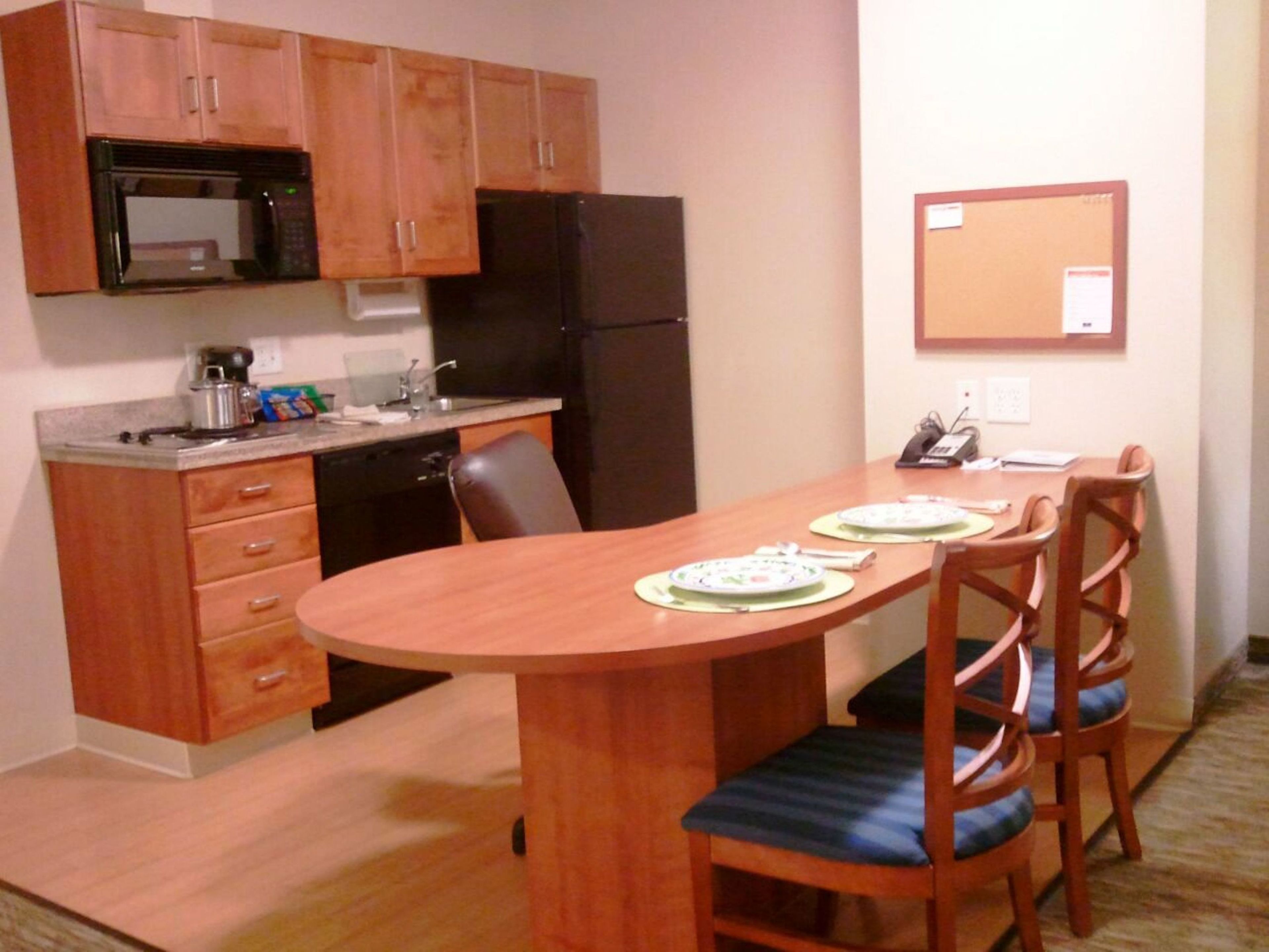 A compact kitchen with wooden cabinets, a black refrigerator, a microwave, and a sink. There is a dining table with two chairs, and a bulletin board on the wall.