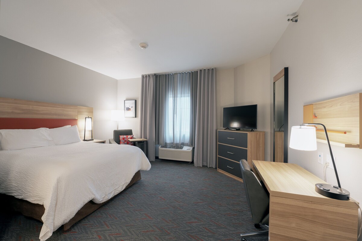 Une chambre d’hôtel avec un grand lit, un bureau avec une chaise, une télévision, un miroir et une fenêtre avec des rideaux.