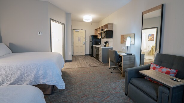 A hotel room with two beds, a desk with a chair, a kitchenette, and a large mirror.