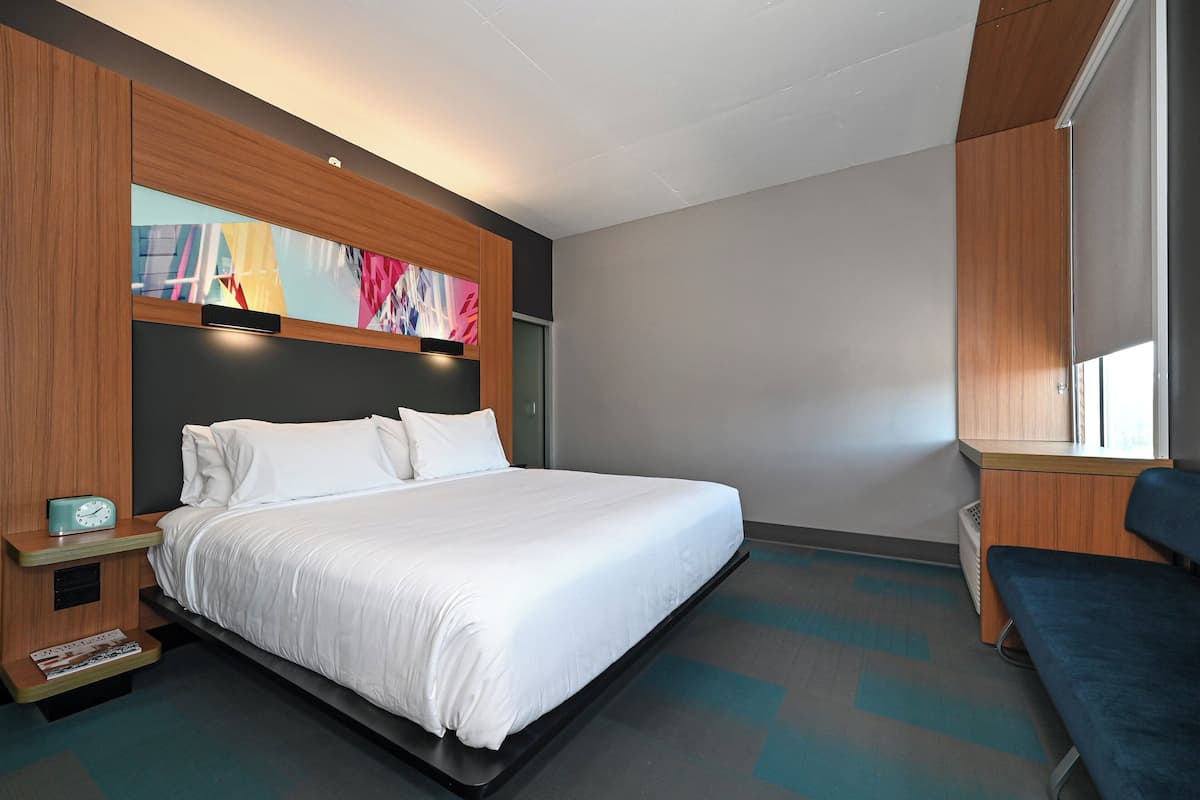 A modern hotel room with a large bed, a wooden headboard with artwork, a bedside table with a clock, and a window with a view.