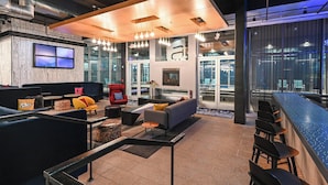 A modern lounge area with seating, a bar, and a large TV screen. - Aloft by Marriott Charleston Airport & Convention Center (North Charleston)