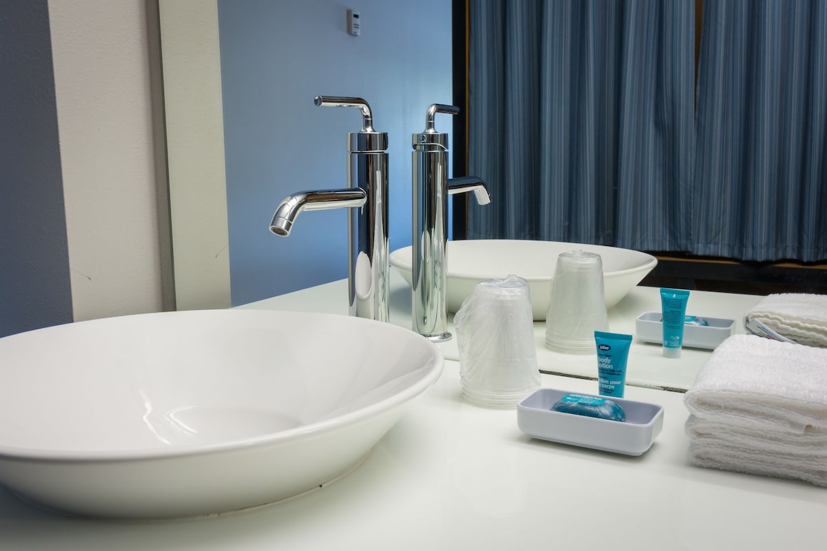 Ein Badezimmer mit einem weißen Waschbecken, zwei chromfarbenen Wasserhähnen, einem Spiegel und Toilettenartikeln auf der Ablagefläche.