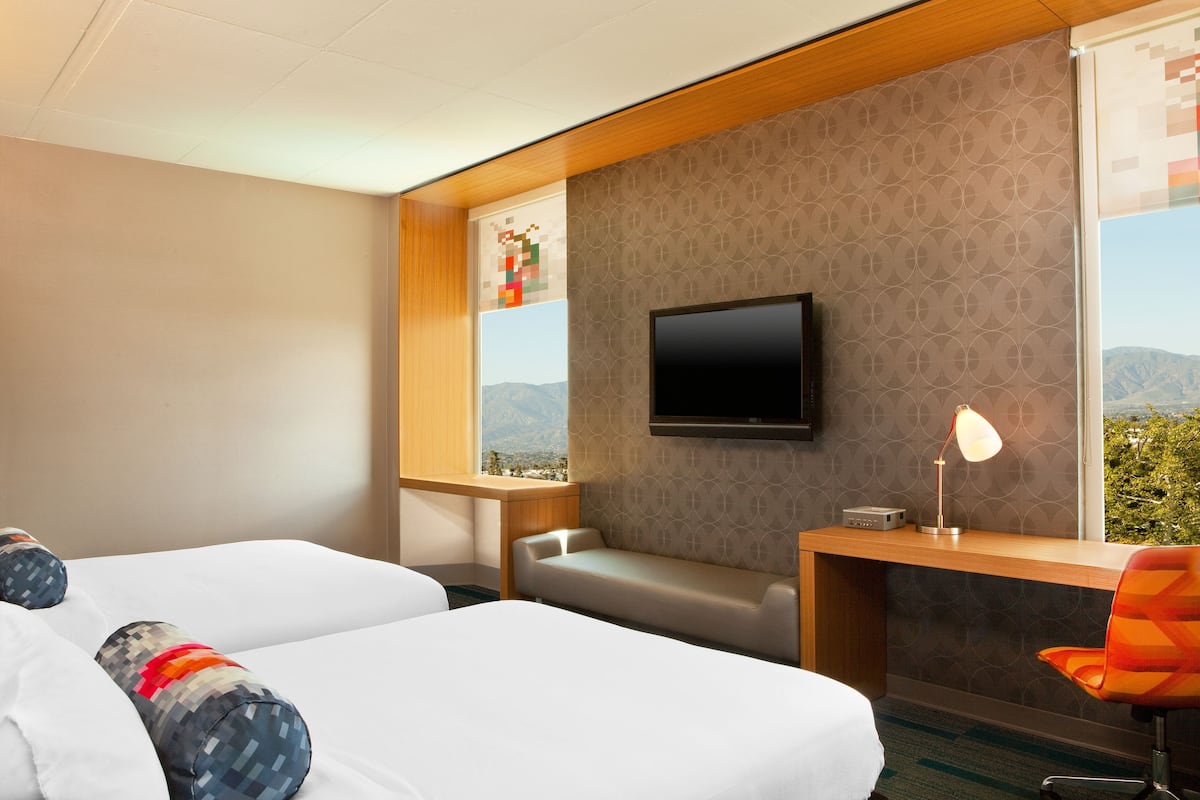 A hotel room with two beds, a desk, a chair, a TV, and a window with a view of mountains.