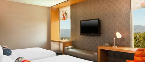 Een hotelkamer met twee bedden, een bureau, een stoel, een televisie en een raam met uitzicht op bergen.