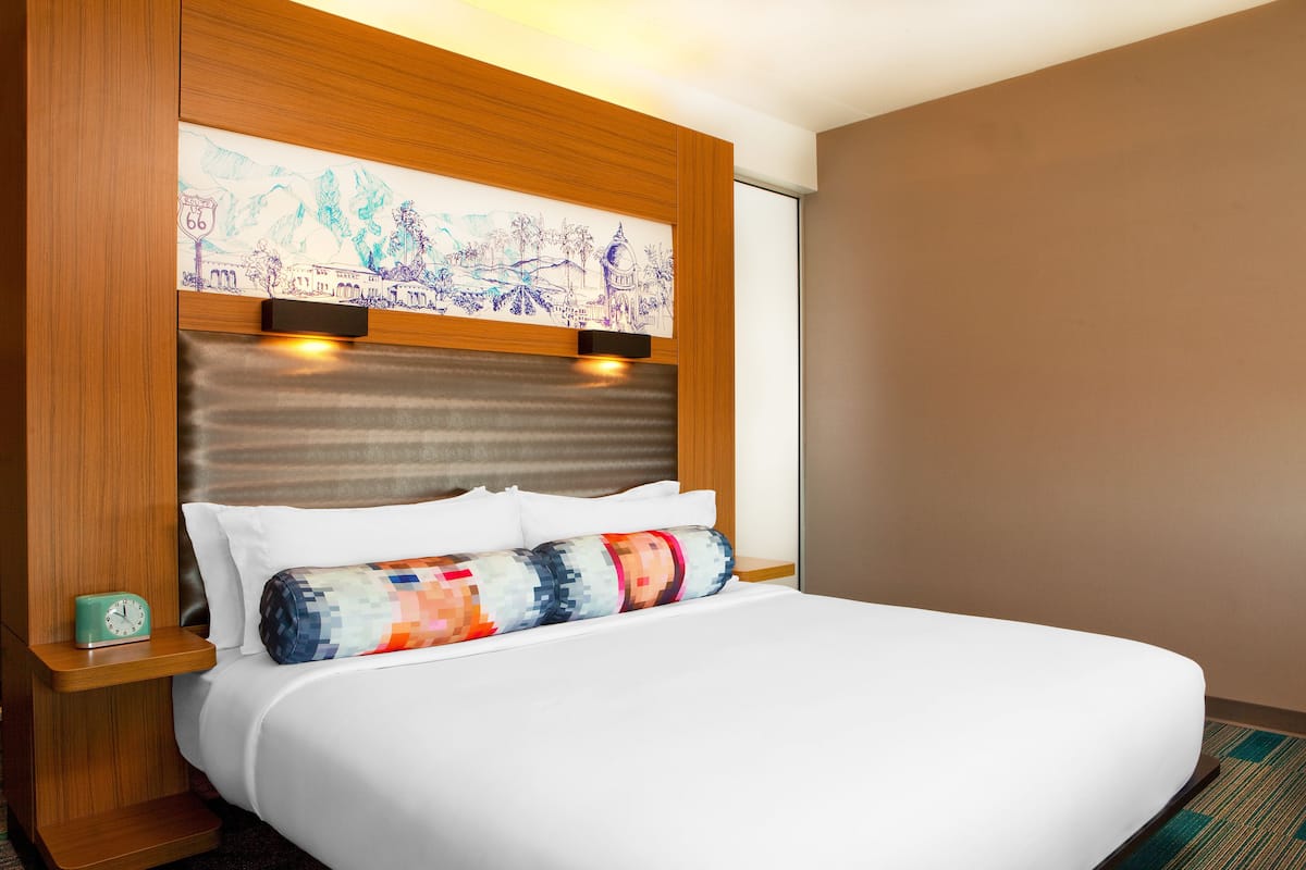 A hotel room with a large bed, a wooden headboard, a wall-mounted artwork, and a small bedside table with a clock.