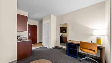 Habitación de hotel compacta con kitchenette, escritorio con silla, espejo grande y una pequeña mesa.