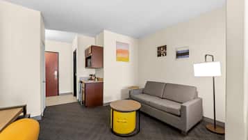 A modern hotel room with a sofa, a small round table, a lamp, a kitchenette, and a door leading to another room.