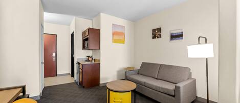 A modern hotel room with a sofa, a small round table, a lamp, a kitchenette, and a door leading to another room.