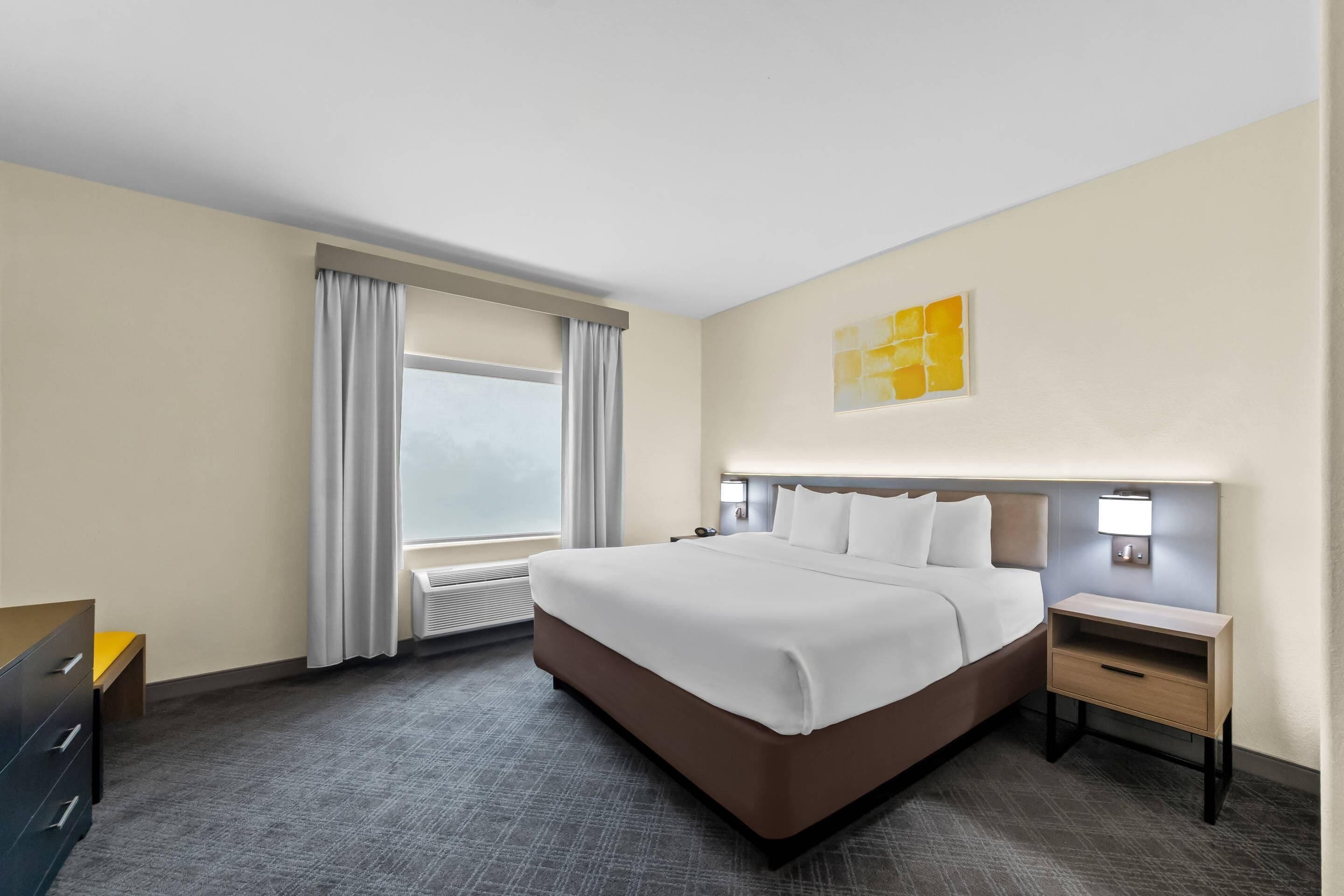Habitación de hotel con una cama grande, una mesita de noche, cortinas en la ventana y una lámpara de pared.