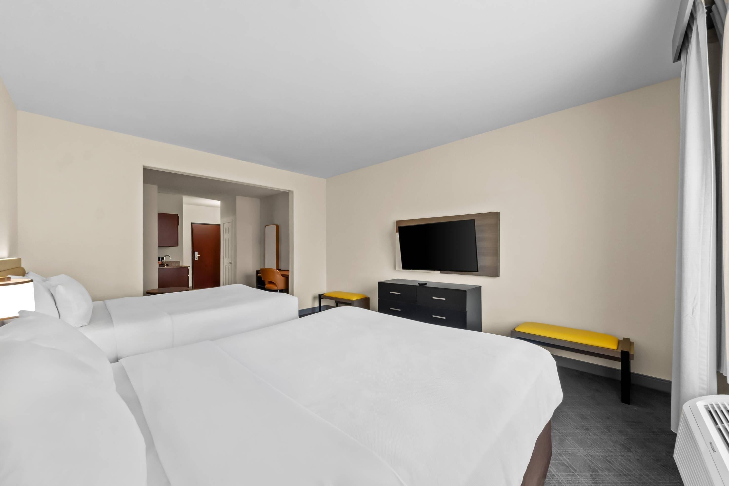 A hotel room with two beds, a TV, a desk with a lamp, a bench, and a door leading to a bathroom.