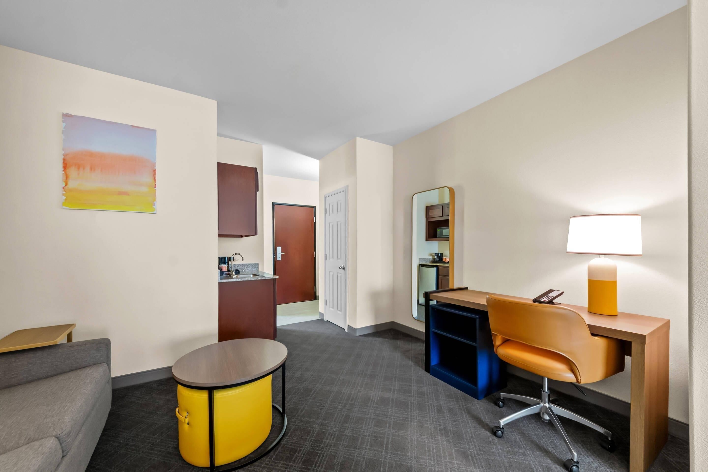 A hotel room with a sofa, a desk, a lamp, a small table, a cabinet, and a door leading to a bathroom.