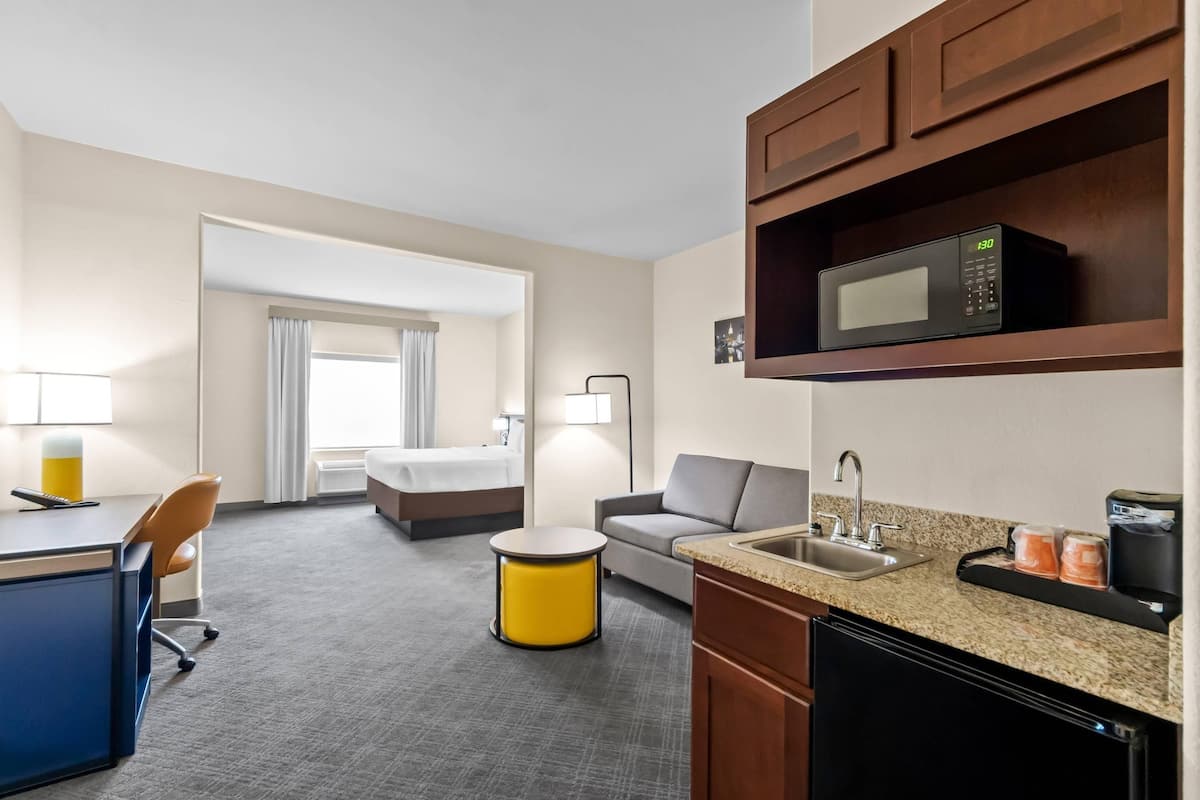 Ein Hotelzimmer mit Küchenzeile, einem Bett, einem Schlafsofa, einem Schreibtisch und einem runden gelben Tisch.