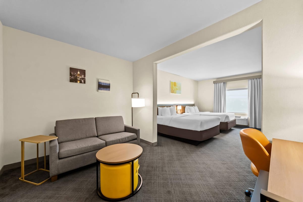 Ein Hotelzimmer mit einer Couch, einem runden Tisch, einem Bett, einer Lampe und einem Fenster mit Vorhängen.