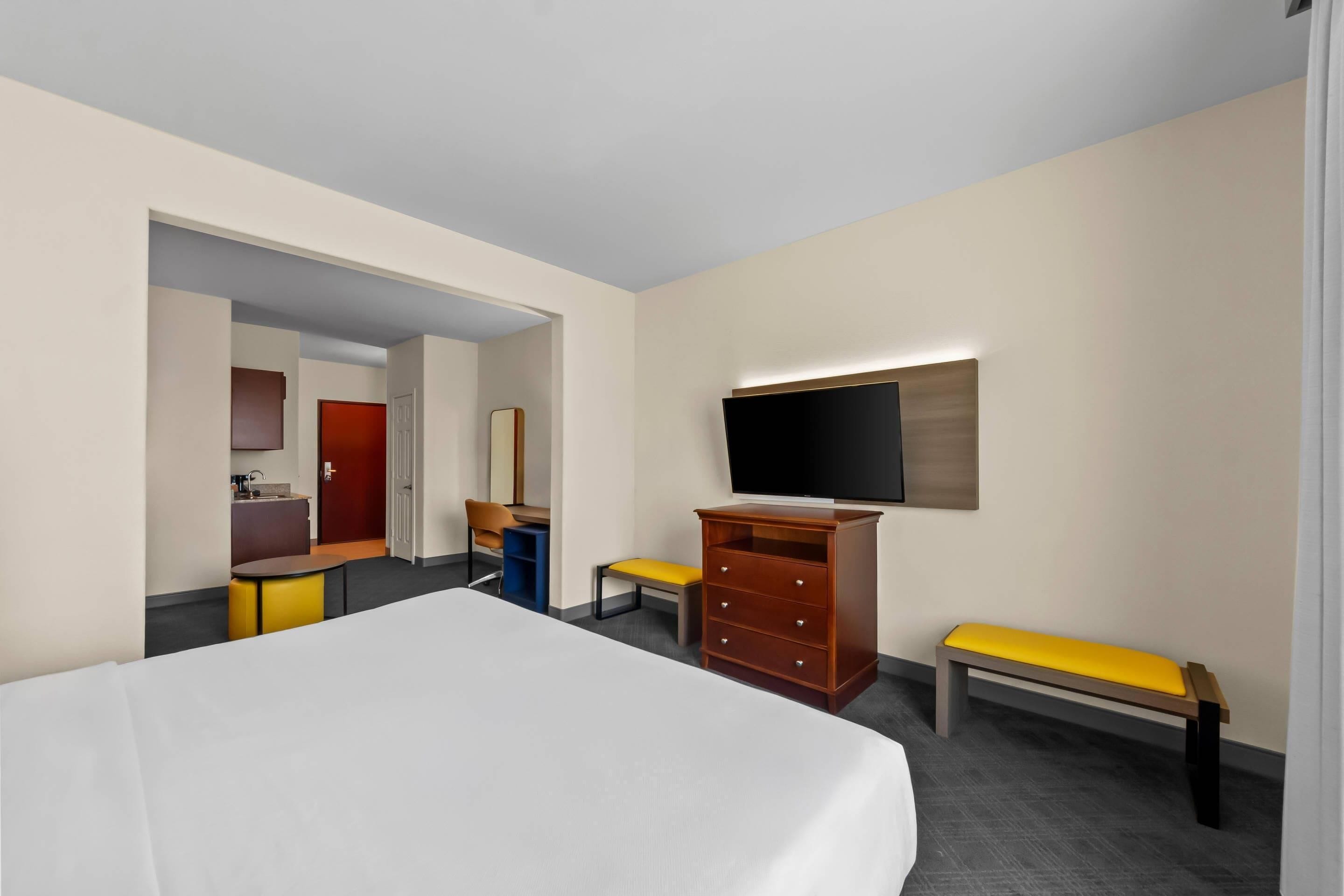 A hotel room with a bed, a TV, a wooden dresser, a bench, and a small table.