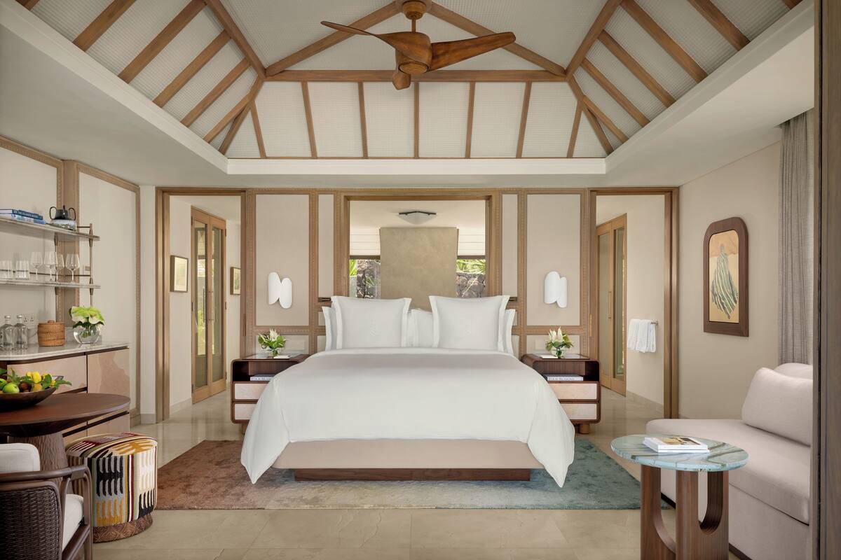 A spacious bedroom with a large bed, wooden ceiling beams, a ceiling fan, bedside tables with lamps, a small round table, and a sofa.