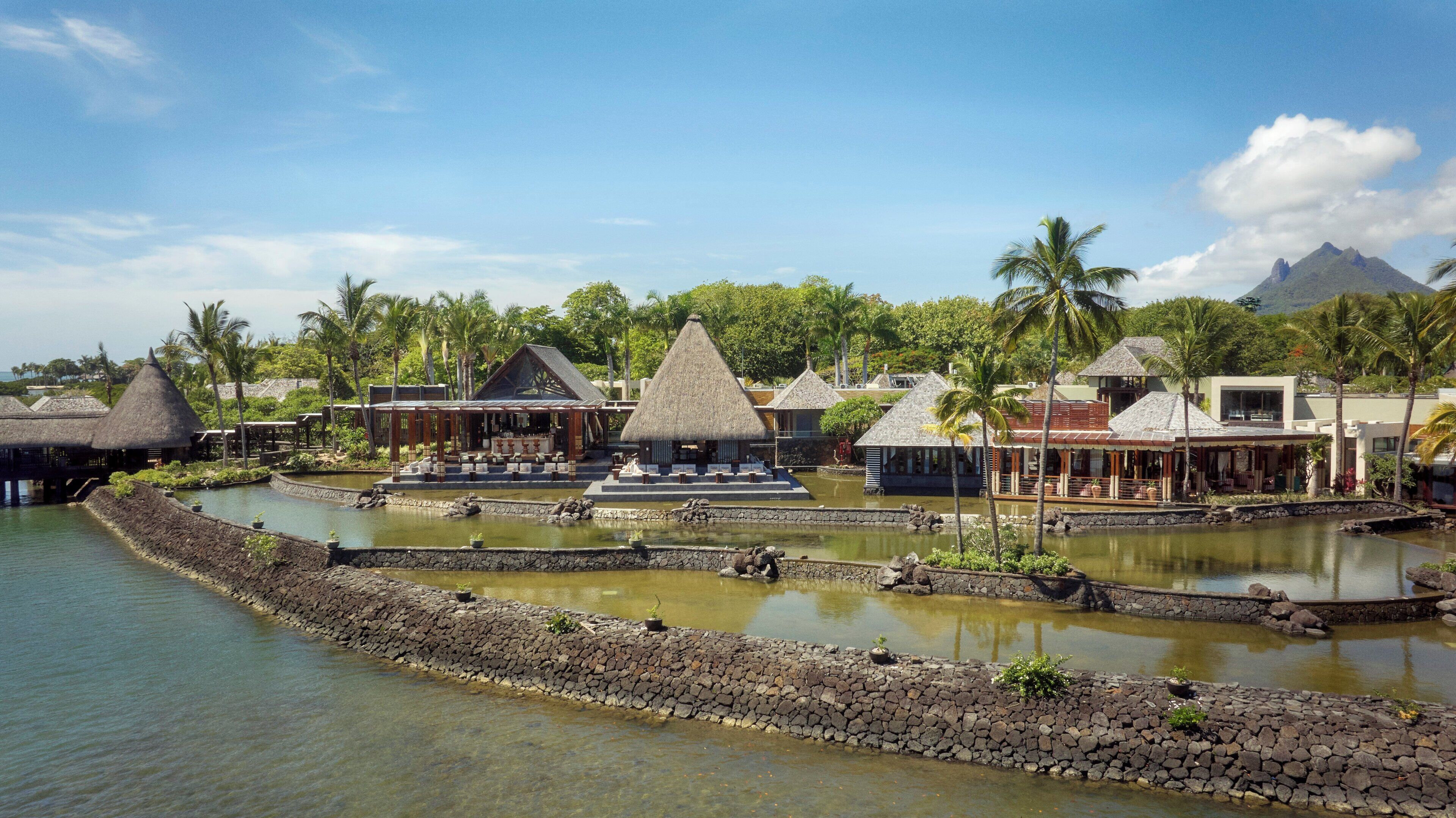 A resort with thatched-roof structures, a central water feature, and a stone wall surrounding a pond.