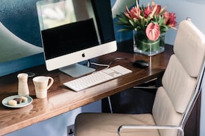 A wooden desk with a computer monitor, keyboard, and mouse. There are two ceramic mugs and a plate with cookies. A vase with flowers is on the desk.