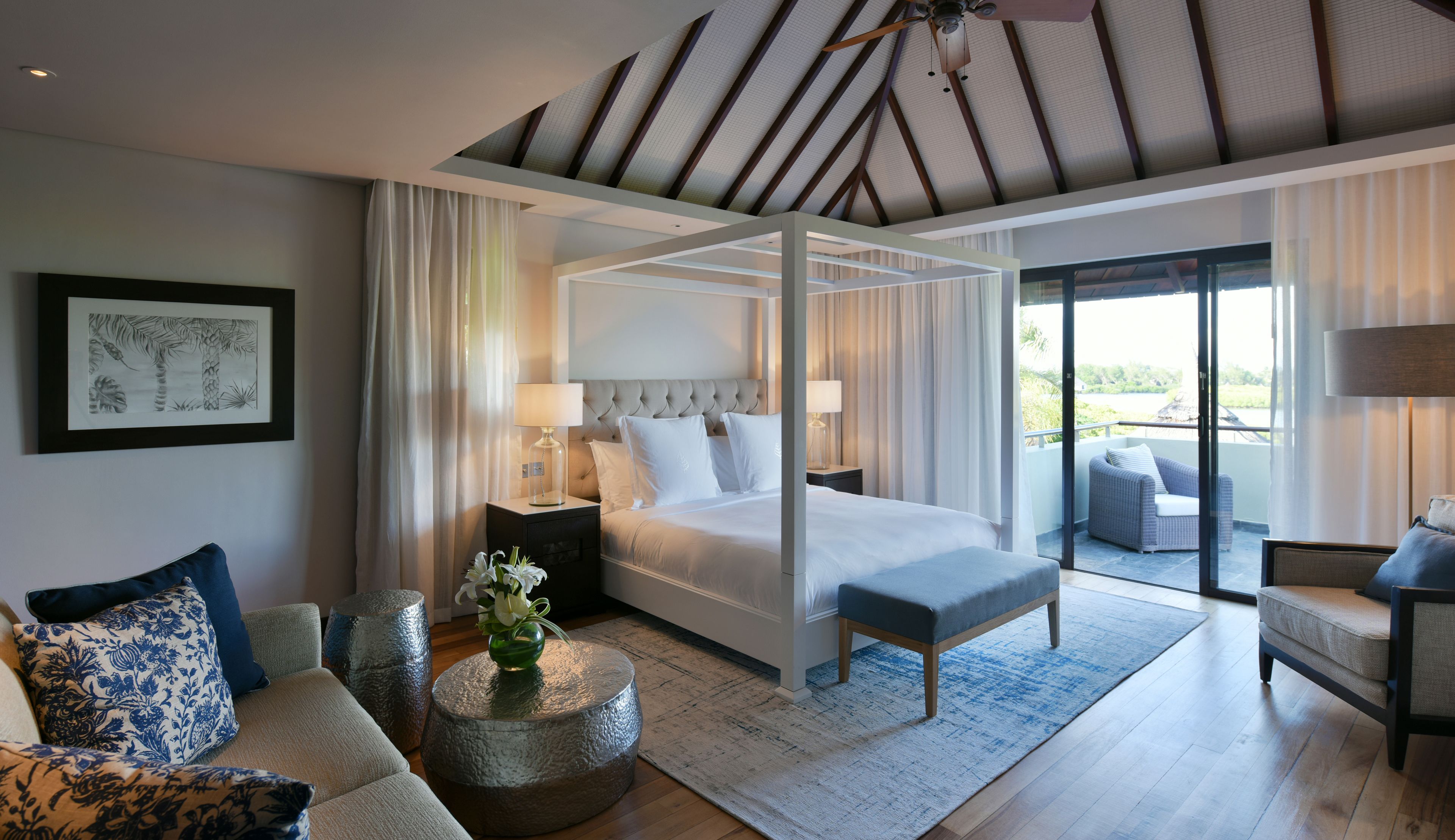 A modern bedroom with a four-poster bed, a sitting area with a sofa and armchairs, a balcony with a view, and a ceiling fan.