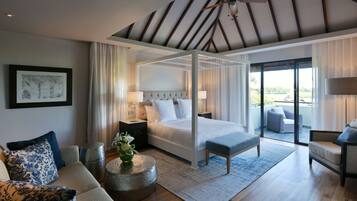 A modern bedroom with a four-poster bed, a sitting area with a sofa and armchairs, a balcony with a view, and a ceiling fan.