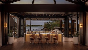 A modern bar area with a high ceiling, wooden beams, and a view of the water. - Four Seasons Mauritius at Anahita (Grand River South East)
