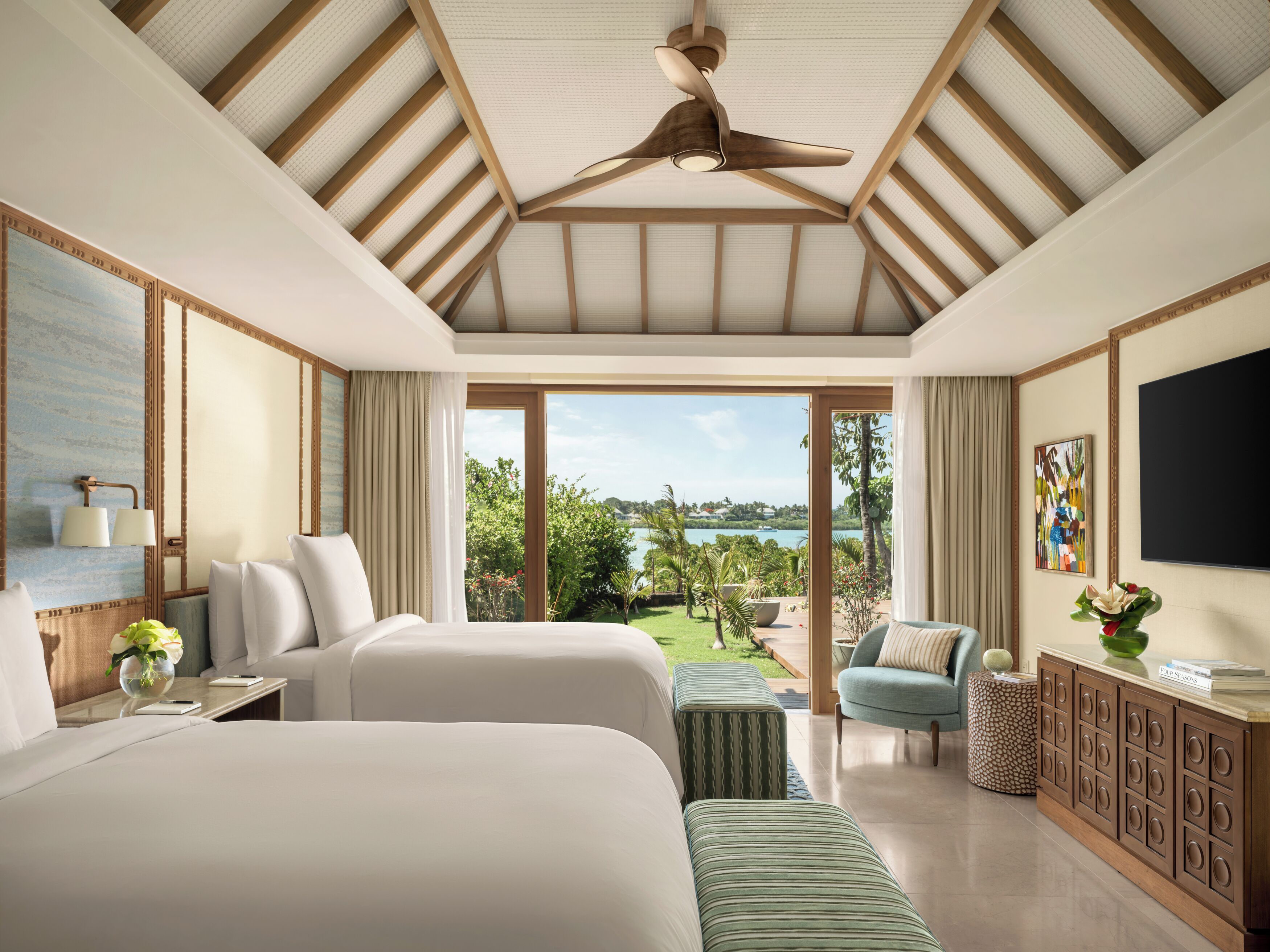 A hotel room with two beds, a ceiling fan, a TV, a view of the outdoors, and a dining area.
