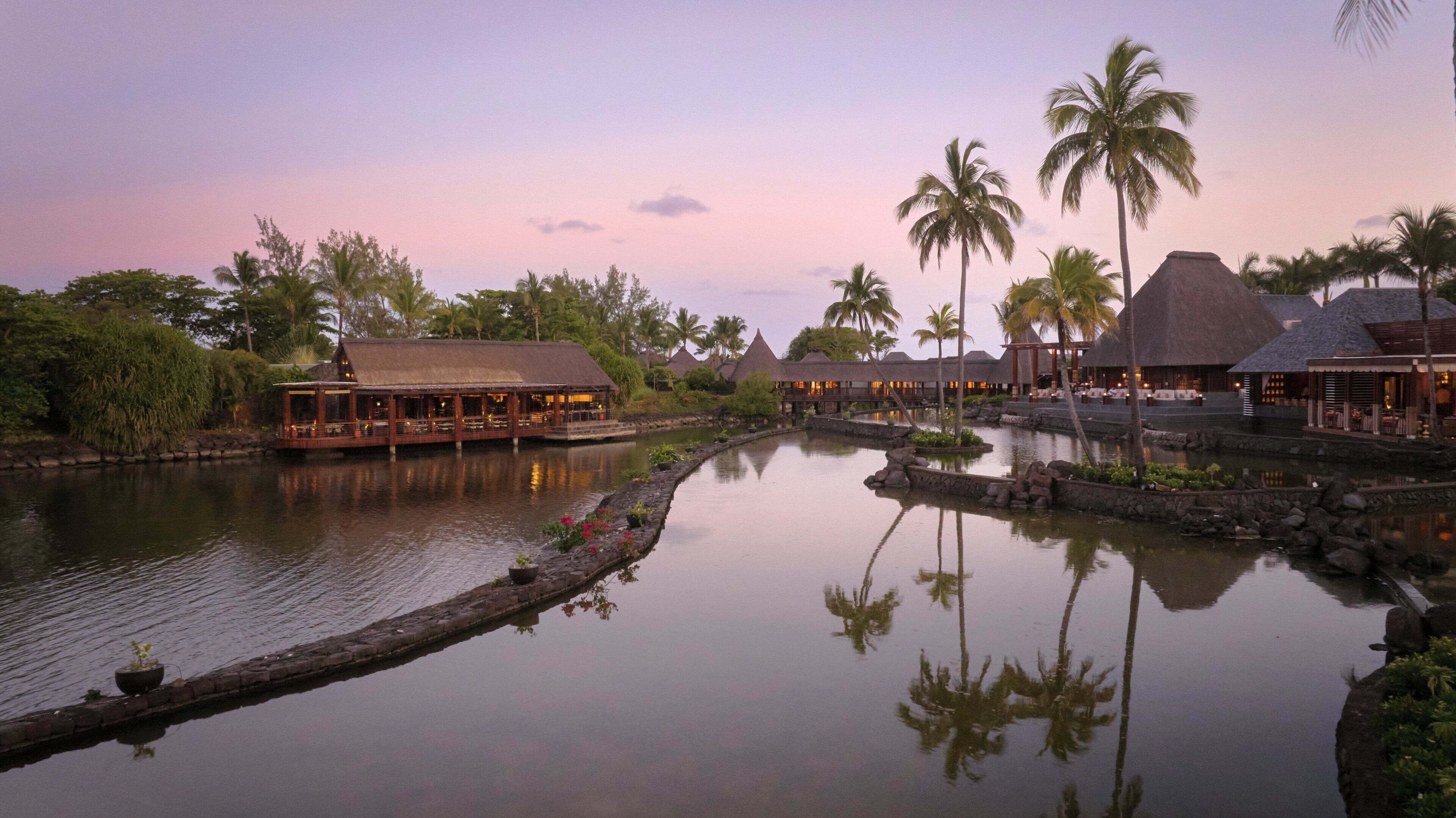 A resort with thatched-roof buildings, a calm water body, and palm trees.
