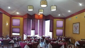 A restaurant with multiple tables and chairs, featuring a red and yellow color scheme, and large windows with purple curtains. - Hotel El Cason and Suites (Chihuahua)