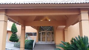 A building entrance with a covered walkway, steps leading to a glass door, and a small garden area with a palm tree. - Hotel El Cason and Suites (Chihuahua)