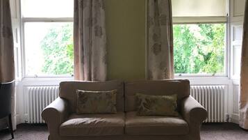 A beige sofa with floral cushions in a room with a large window, curtains, and a radiator.