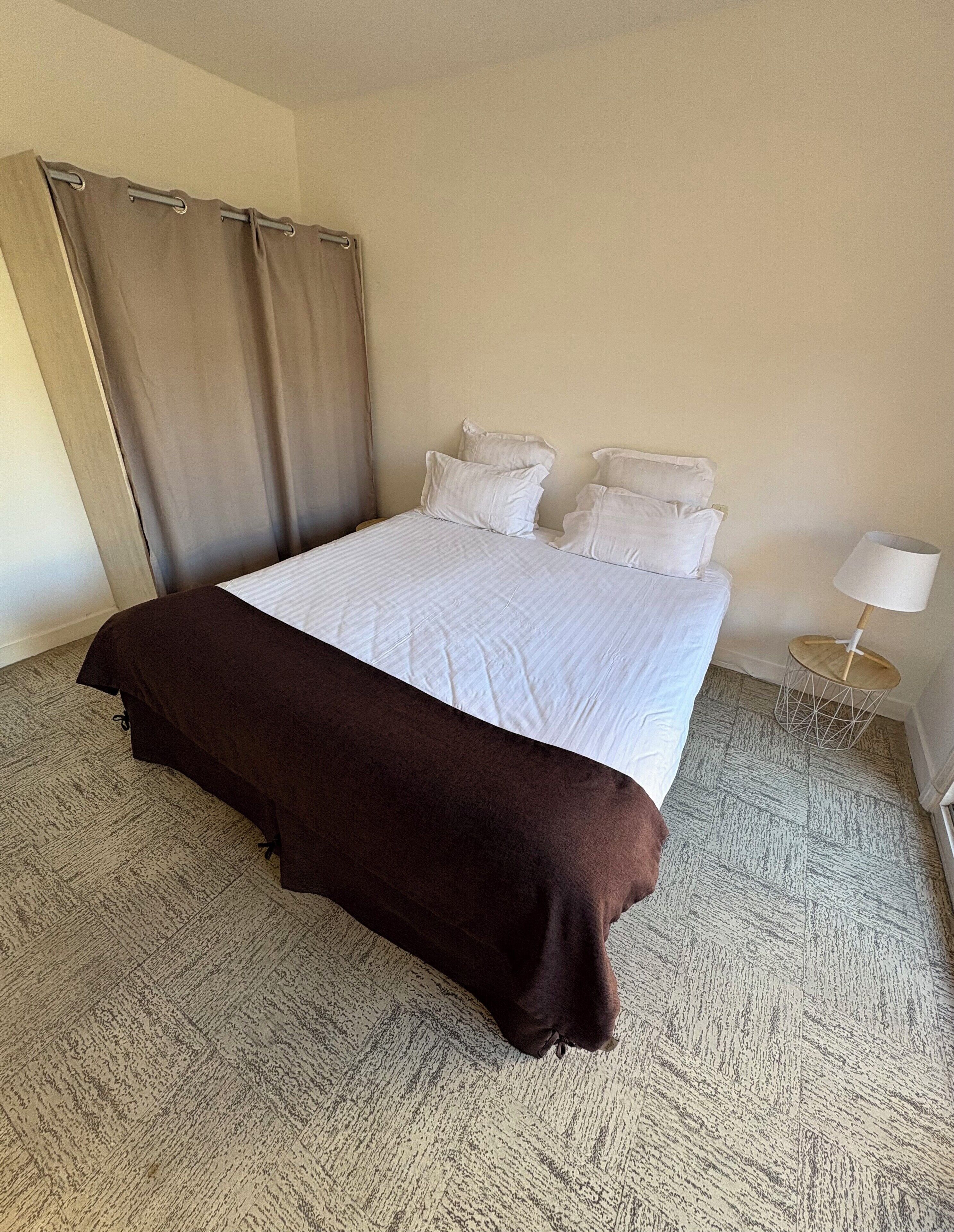 A hotel room with a large bed, a brown throw blanket, a side table with a lamp, and a curtain.