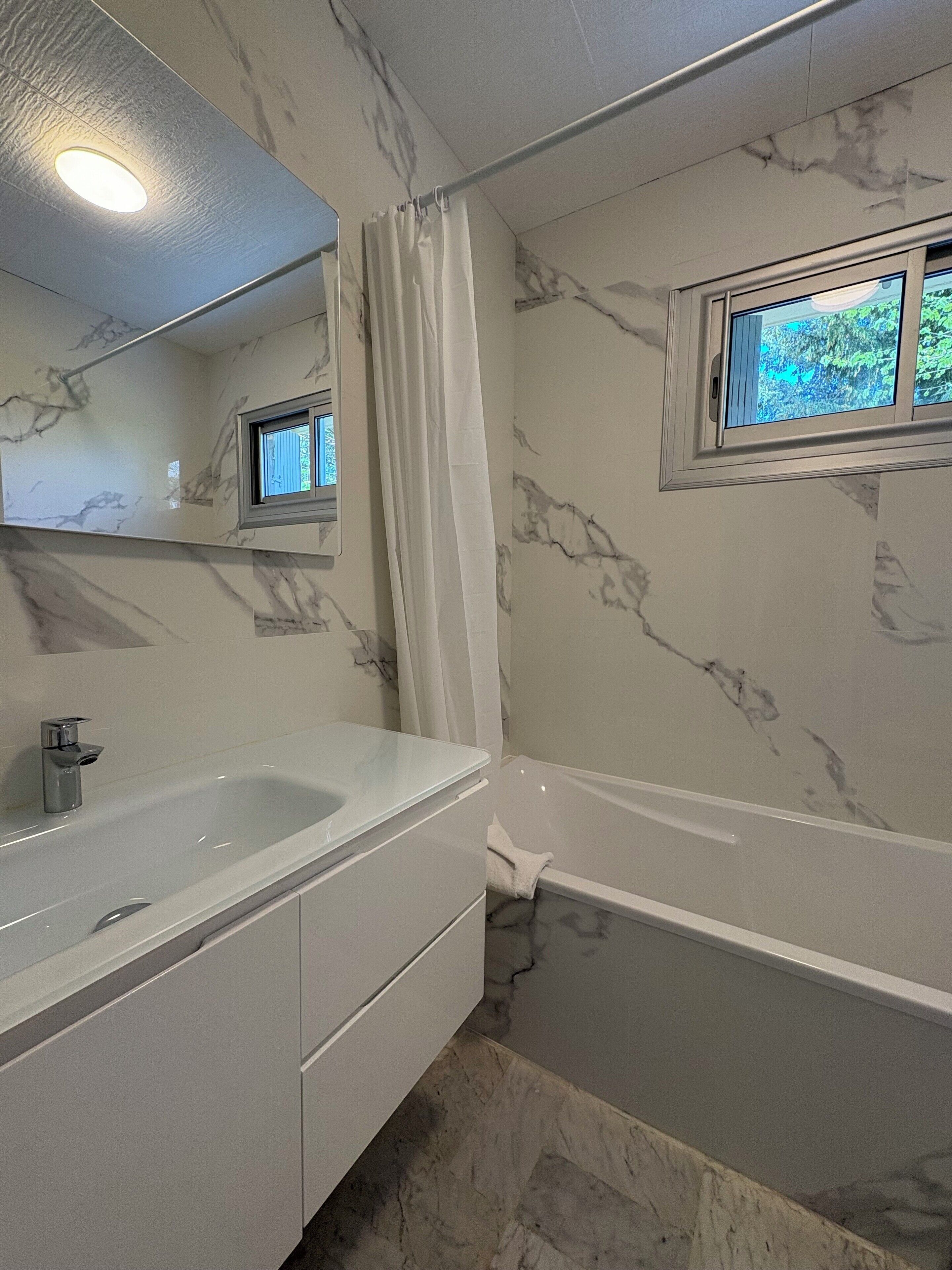 A bathroom with a marble wall, a bathtub, a sink with a mirror, and a window with a view of greenery.