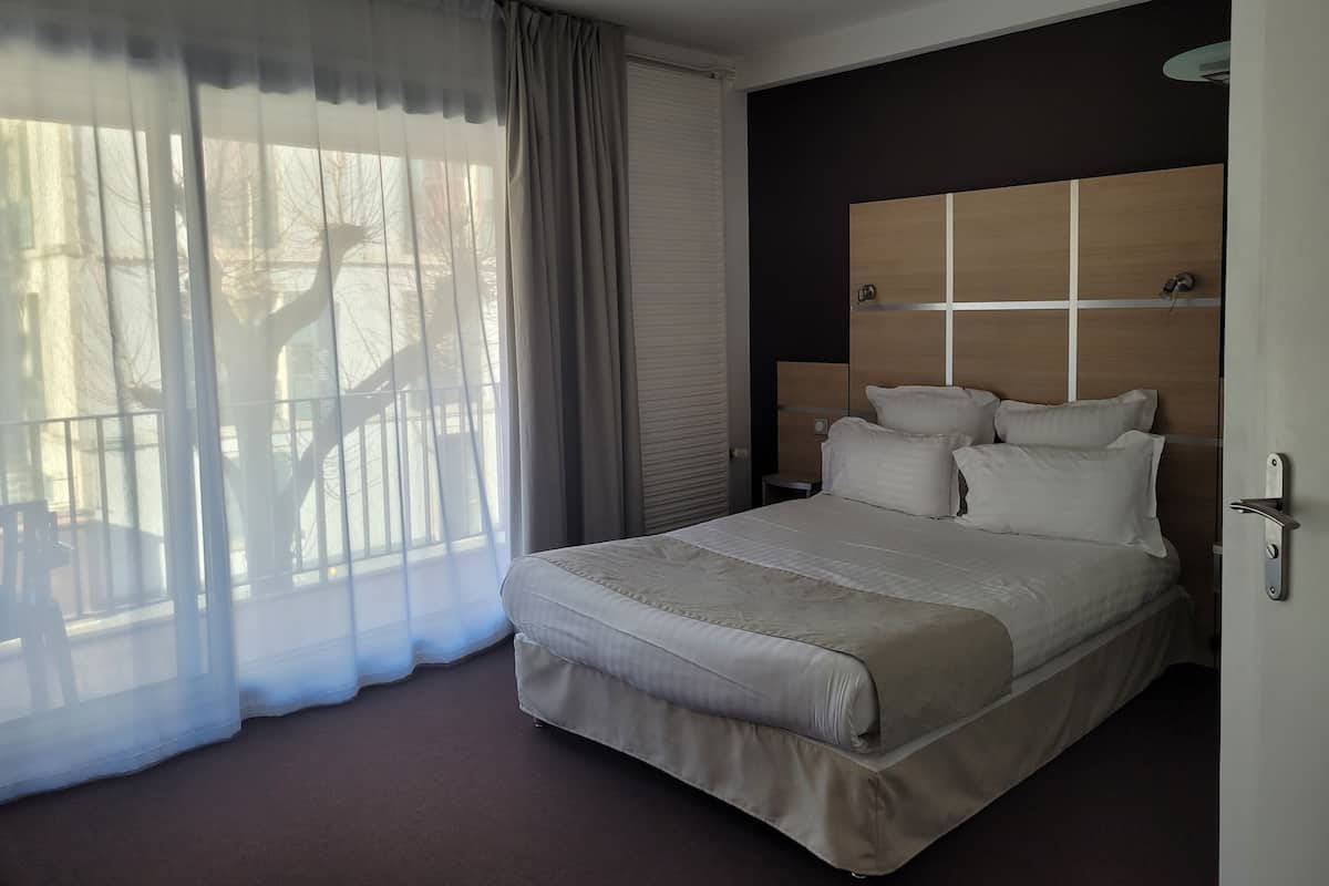Una cama bien hecha con cabecera de madera, un ventanal con cortinas translúcidas y vistas a un balcón.