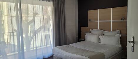 Una cama bien hecha con cabecera de madera, un ventanal con cortinas translúcidas y vistas a un balcón.