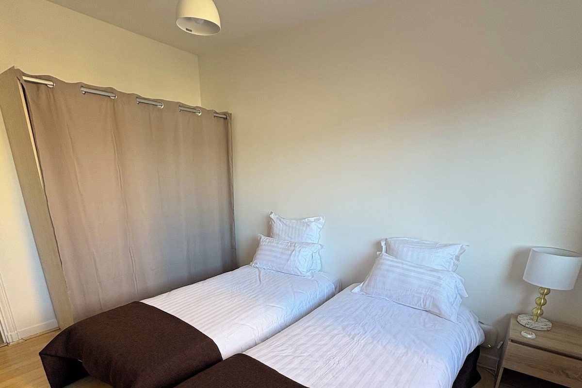 Un dormitorio con dos camas, piso de madera, una cortina, mesita de noche con lámpara y una lámpara colgante.