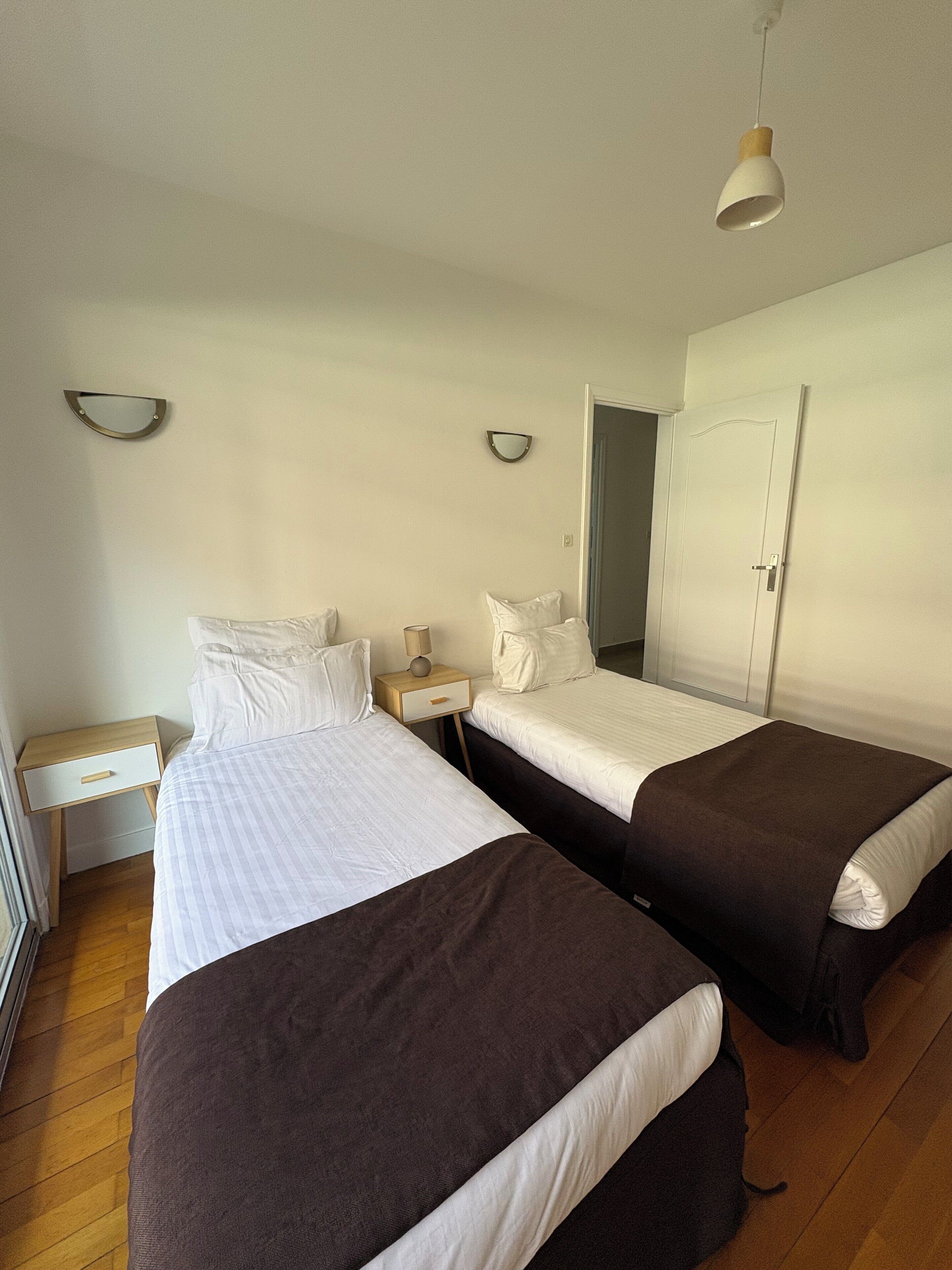A hotel room with two beds, a bedside table with a lamp, a mirror, and a door.