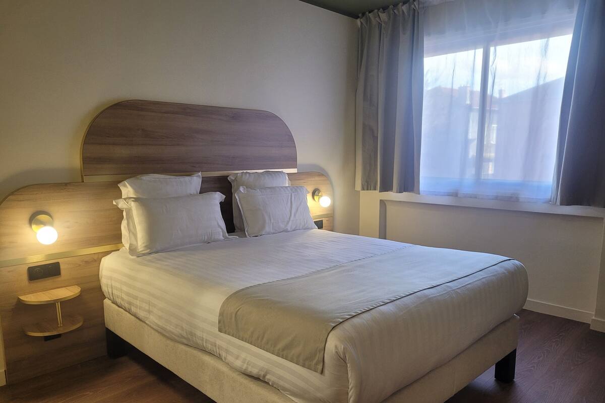 Una cama bien hecha con cabecera de madera, dos lámparas de noche y una ventana con cortinas.