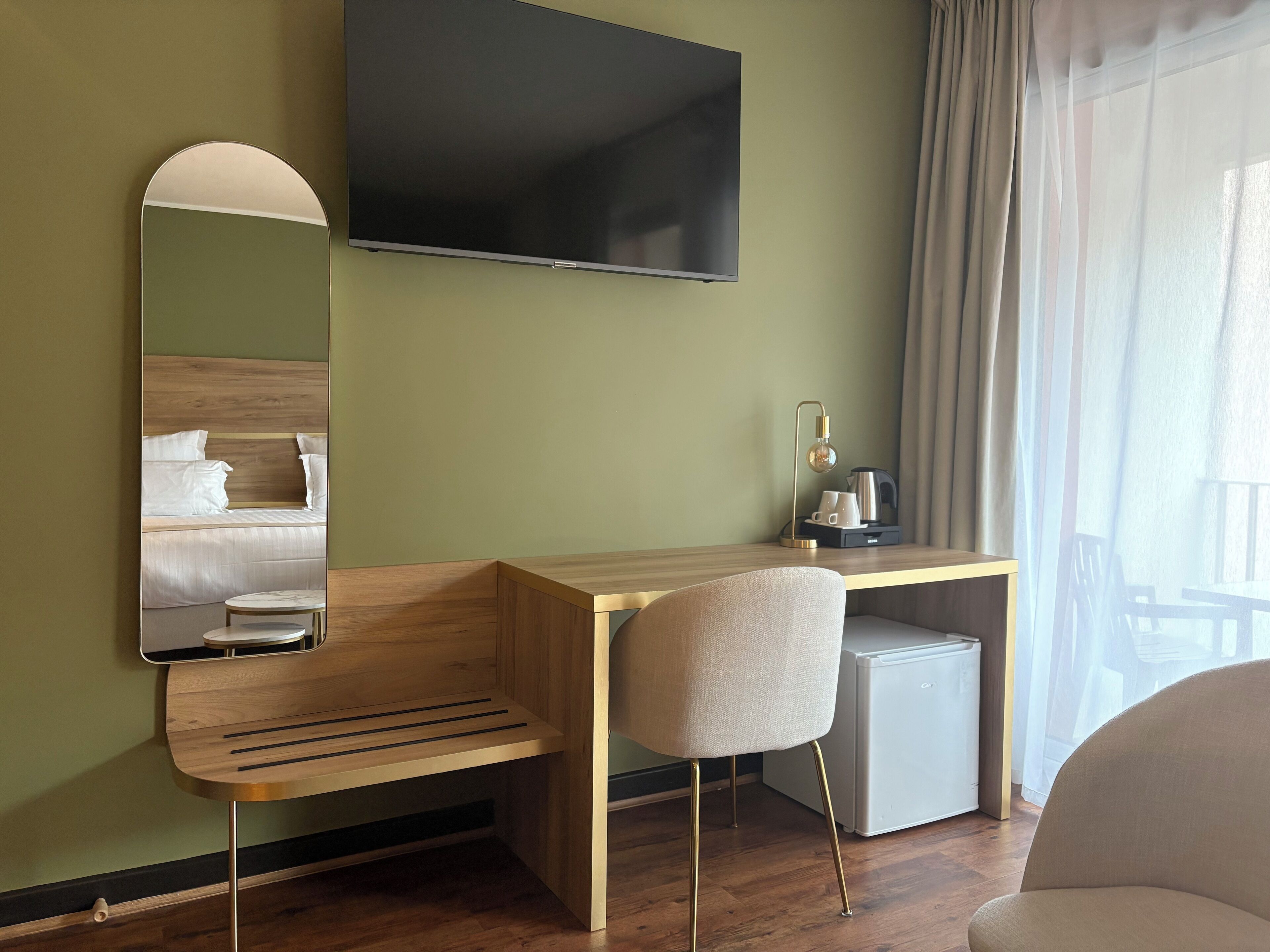 A hotel room with a wooden desk, a small refrigerator, a television, a mirror, and a chair.