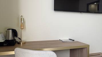 Una habitación de hotel moderna con un escritorio de madera, una silla blanca, un televisor de pantalla plana montado en la pared y una cafetera sobre el escritorio.